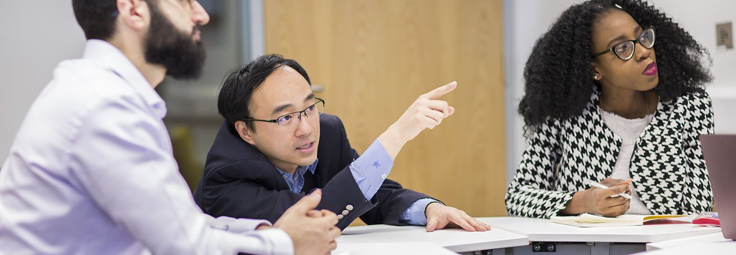 Professor Yang at desk with students pointing at the board and explaining something.