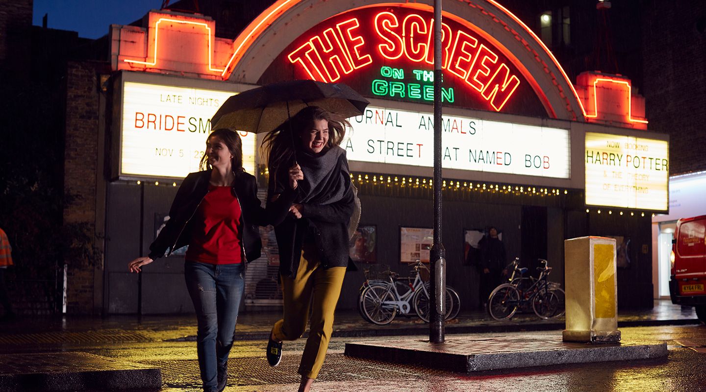 Two young females crossing the road with open umbrella outside of the screen on the green cinema