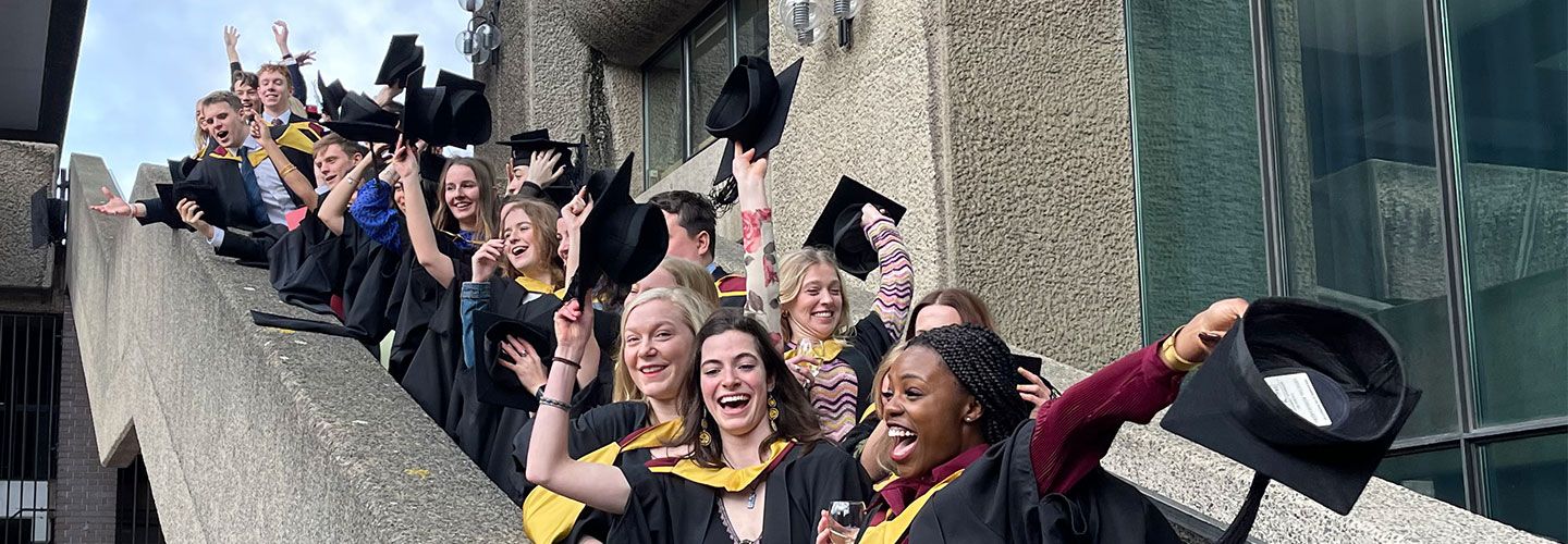 Students stand on the steps leading to the Barbican Centre in their graduation gowns and throw their mortarboards into the air
