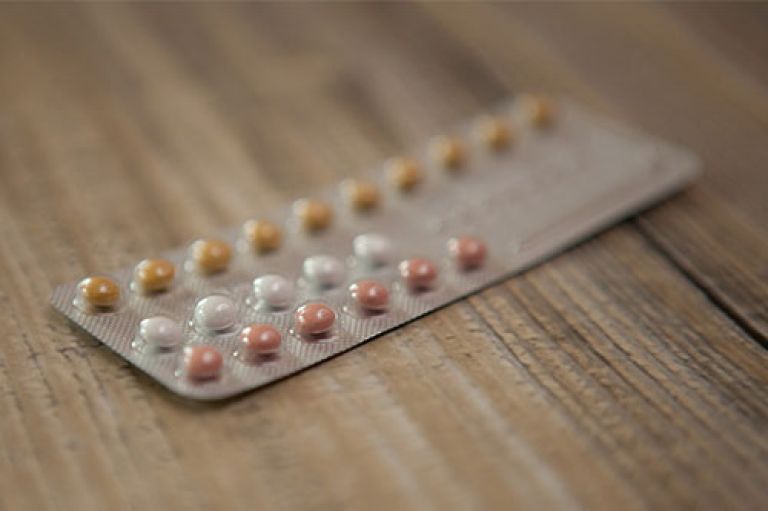 Image of a pack of contraceptive pills (a row of yellow, then a row of white, then a row of pink pills) against a wooden table