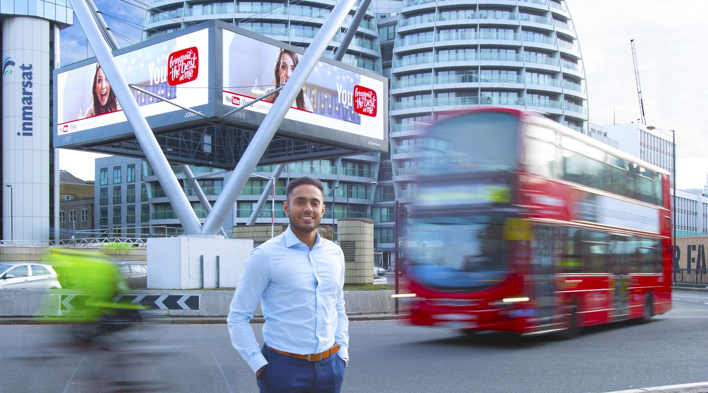Student smiling outside Old Street roundabout with red double decker bus driving behind