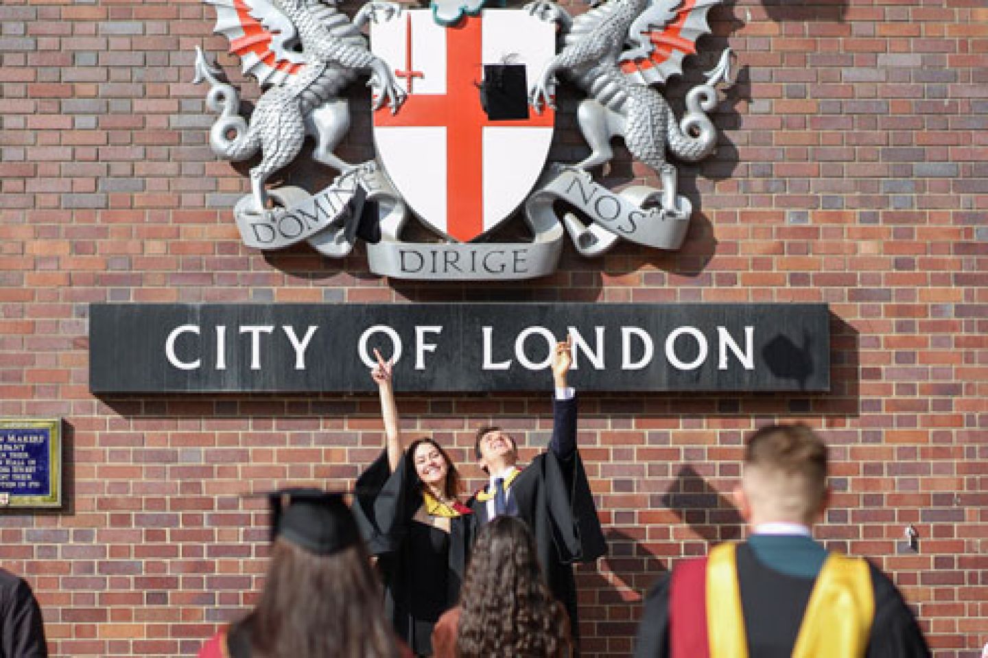 Two graduates throw their hats in the air in front of the City of London sign.