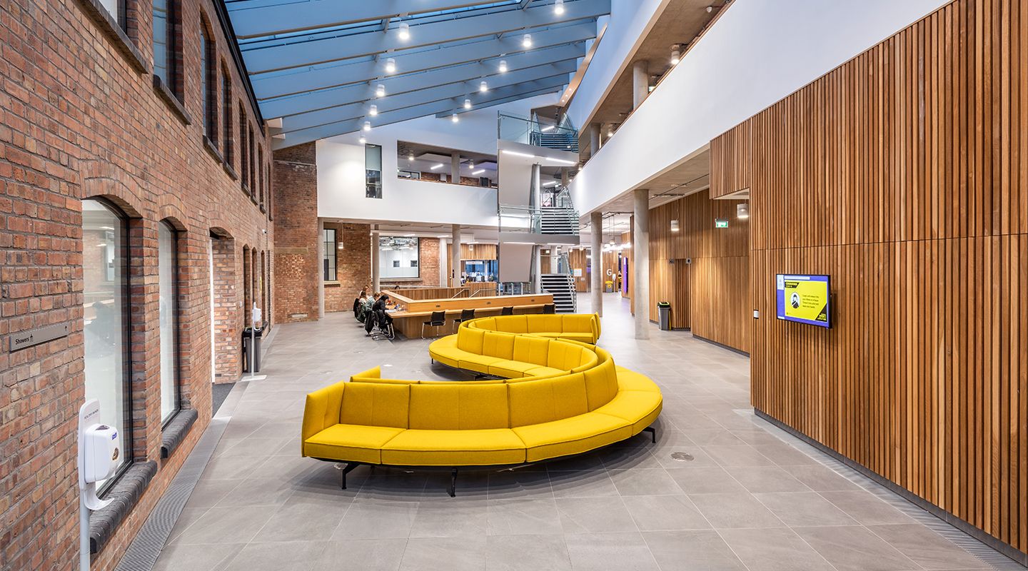 Large yellow curved sofa centred in an open space foyer with students working at desks in the background