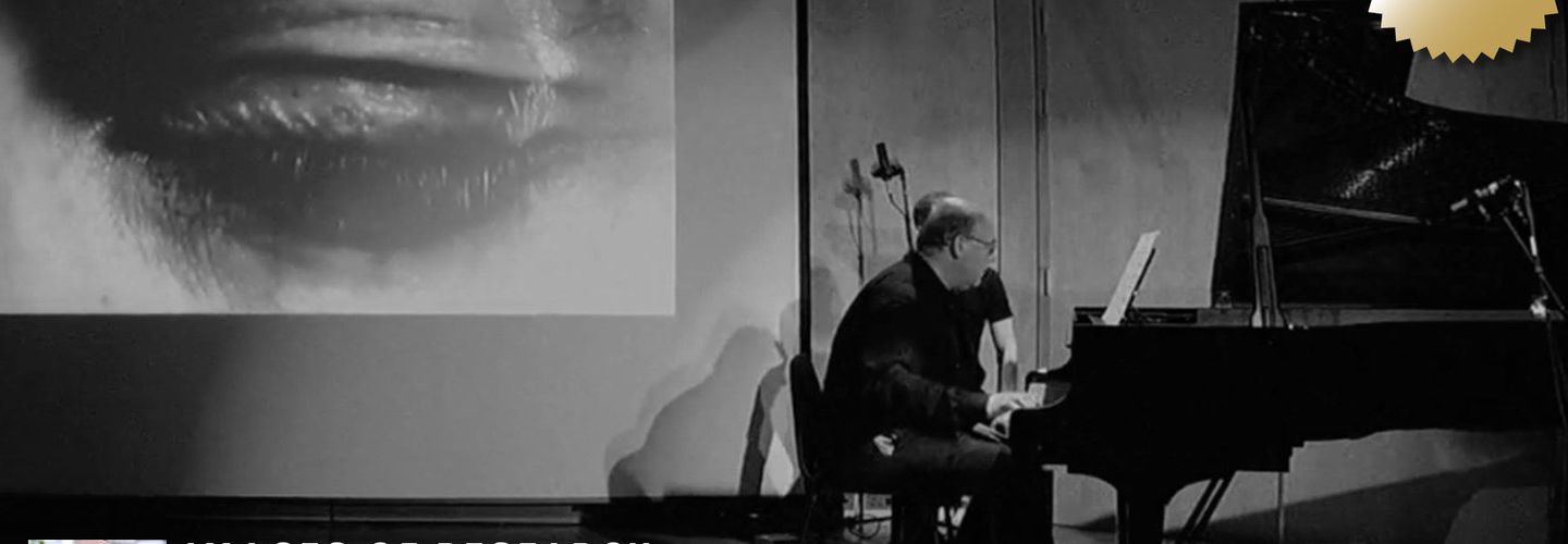 Dr Ian Pace playing a piano on stage in front of a large screen showing a projection of a closed human eye.