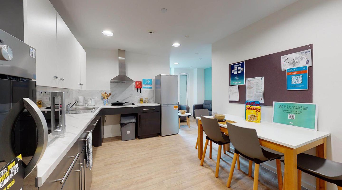 Large kitchen with oven, hob, microwave, sink, fridge-freezer and breakfast bar against the wall. Small lounge with sofas at the far end. Black and white cabinets, pale wood floor and notice board above the breakfast bar.