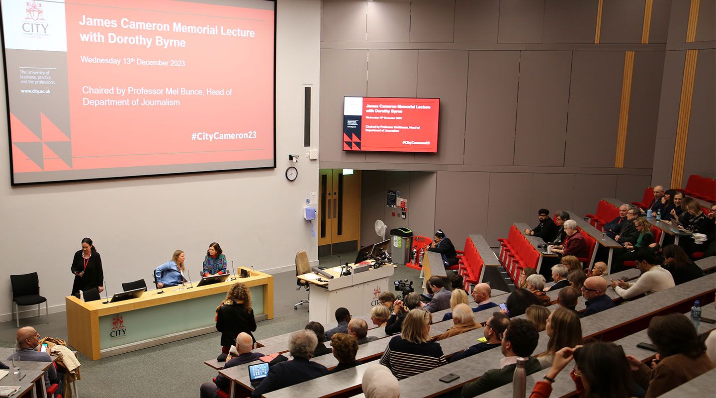 Journalist Dorothy Byrne and Prof Mel Bunce sit in front of a desk in a large auditorium full of people.