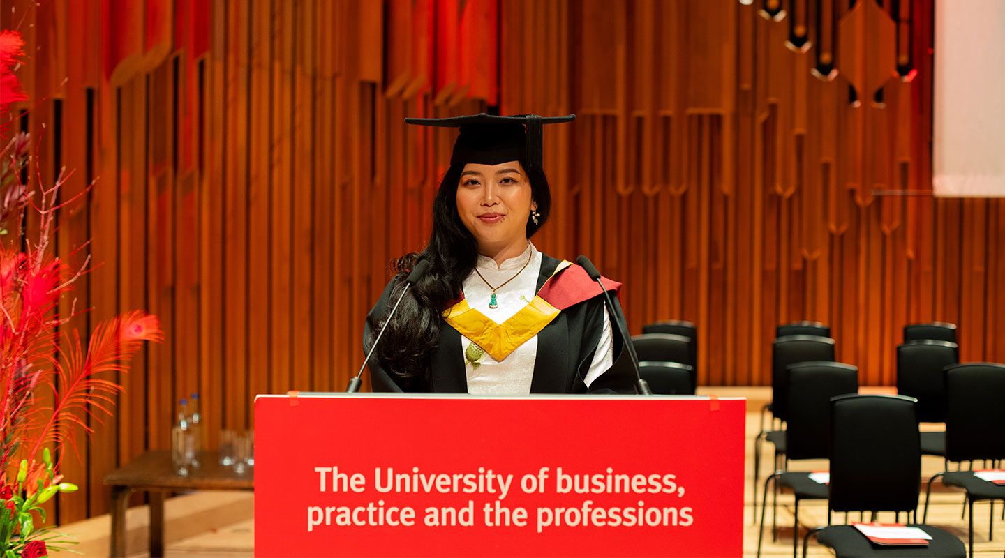 Picture of Sally Mieu, wearing her mortarboard and gown, smiling into the camera. She is wearing a jade necklace and earrings and a smart, white shirt. She is standing in front of a lectern with the word 'University of business, practice and the professions' printed in white against a red background. Behind her are red plants and the wooden walls of the graduation hall in the Barbican Centre
