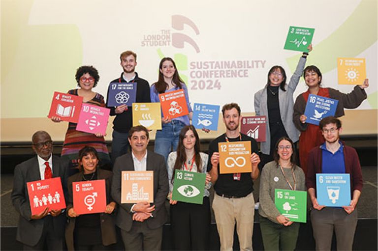 Image of two rows of people standing and smiling to camera. Behind them is a screen that reads The London Student  Sustainability Conference 2024. Each person holds a brightly coloured square tile with a number and infographic on it, referencing one of the 17 UN Global Development Goals