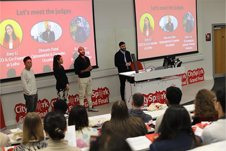 Image of the judges standing in front of an amphitheatre of people. Behind them are red screens with the words CitySpark