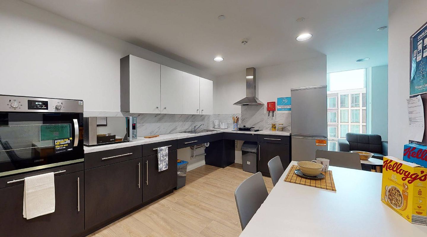 Large kitchen with oven, hob, sink, fridge-freezer, cupboards and breakfast bar against the wall. Black and white cabinets, white walls with grey marble effect tiles over the work surface, pale wood floor and small lounge with sofas at the far end with large windows.