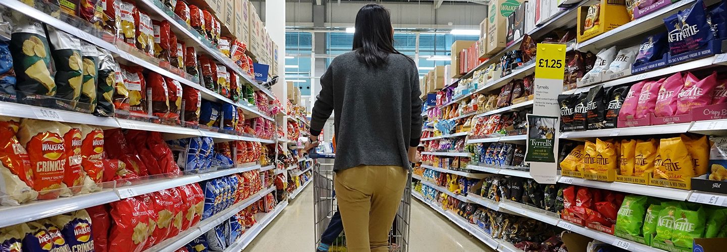 Woman in a supermarket pushing a trolley past rows of crisps.