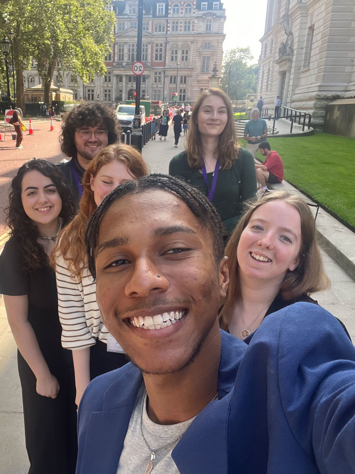 Student Miles Johnson smiles to the camera. He is taking a selfie and behind stand a group of fellow students also smiling. They are standing in West London and there are big buildings behind them, grass to one side and and trees to the other.