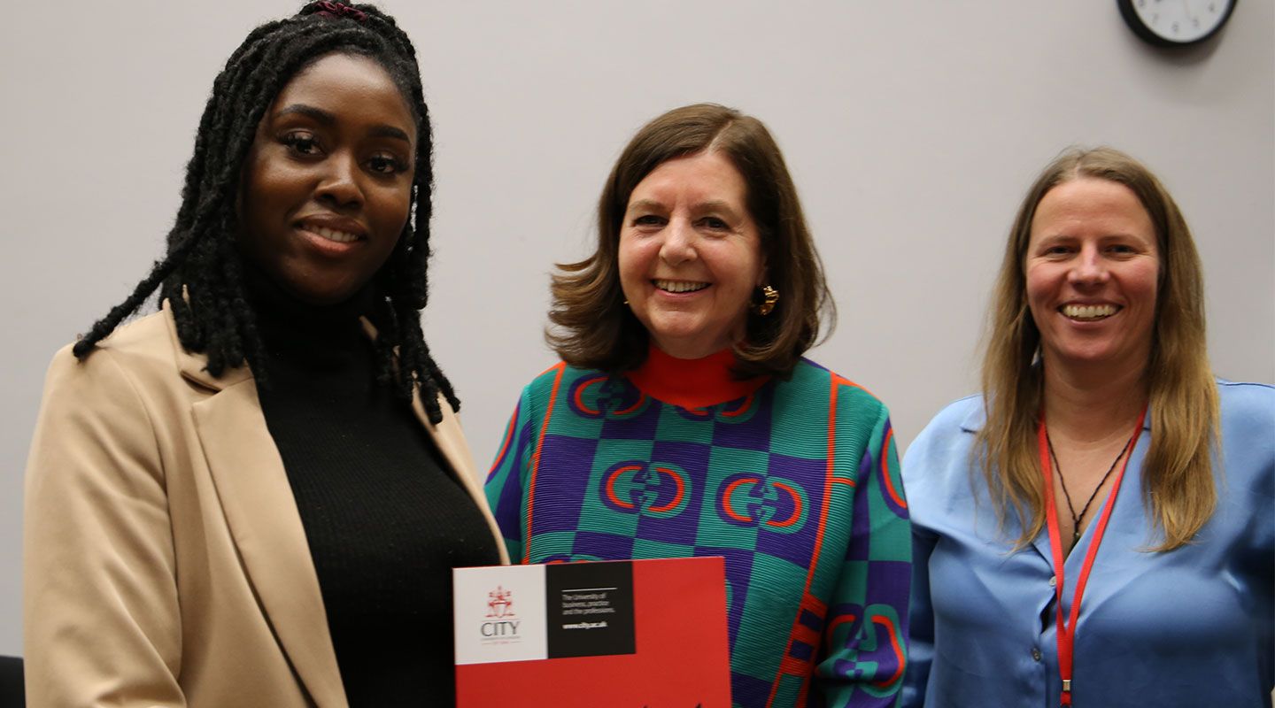 Runako Celina (left) holds her Eric Robins Prize award (a black and red patterned card) with journalist Dorothy Byrne (centre), next to Prof Mel Bunce (right)
