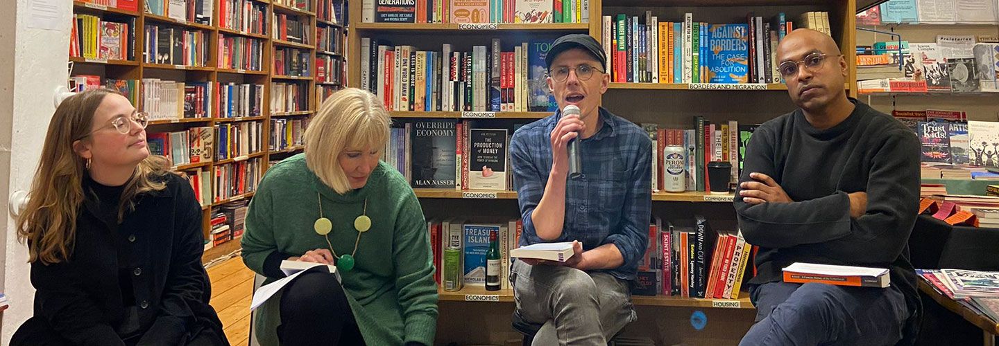 A landscape image of four speakers sitting and facing the camera at the book launch of Dr Dom Davies' book. Behind them are row of colourful books on bookcases. There are two women to Dom's left and one man to his right.