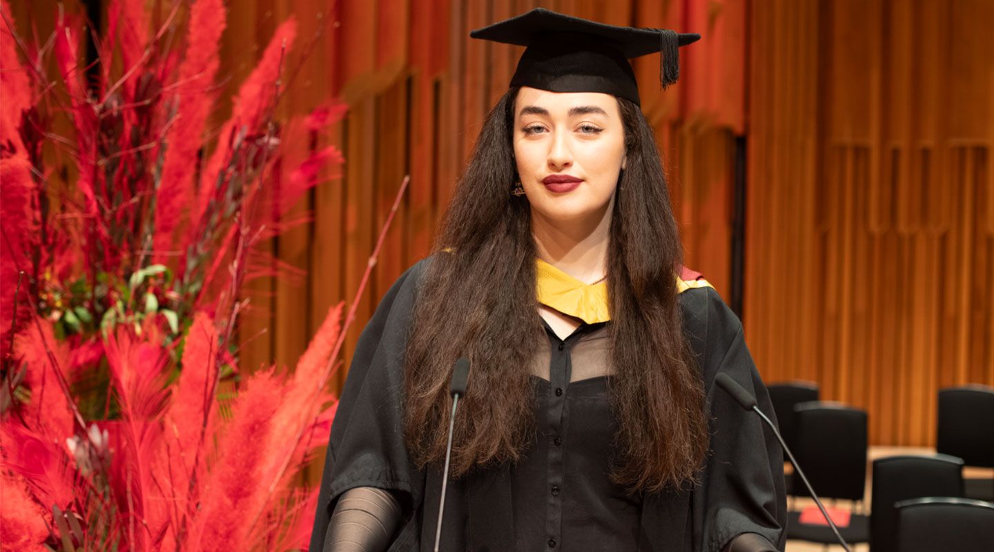 Student speaker Vanessa Igoe (MA Broadcast Journalism) look towards the camera, wearing her mortarboard and robe. To her left are red plants, to her right, seats. Behind her in the wooden structure of the Barbican Hall in which she graduated.