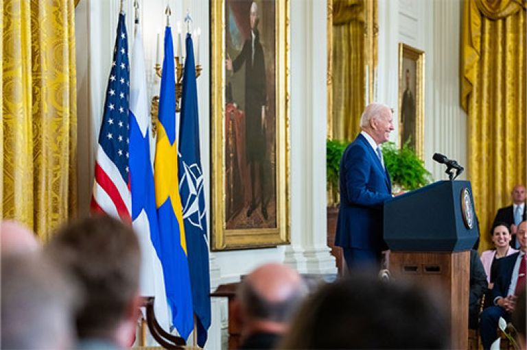 Image of US President Biden speaking at an official event. He smiles and stands in front of a lectern with a microphone. Around him is a crowd of people sitting. To one side is the American flag.