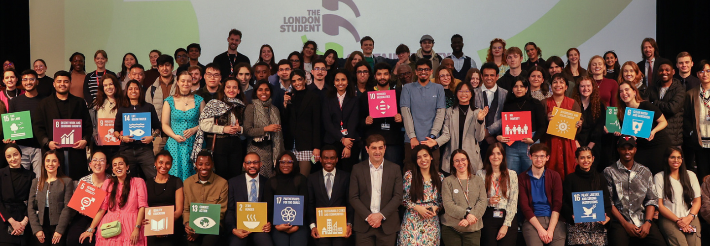 Image of three to four rows of people standing and smiling to camera. Behind them is a screen that reads The London Student  Sustainability Conference 2024. Each person holds a brightly coloured square tile with a number and infographic on it, referencing one of the 17 UN Global Development Goals