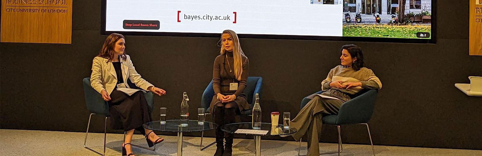 Image of speaker Natalie Bonnick, Dr Stephanie A Baker and Daniella Isaacs sitting on chairs on stage at Bayes Business School in an amphitheatre lecture room. Behind them is a large screen with the event title