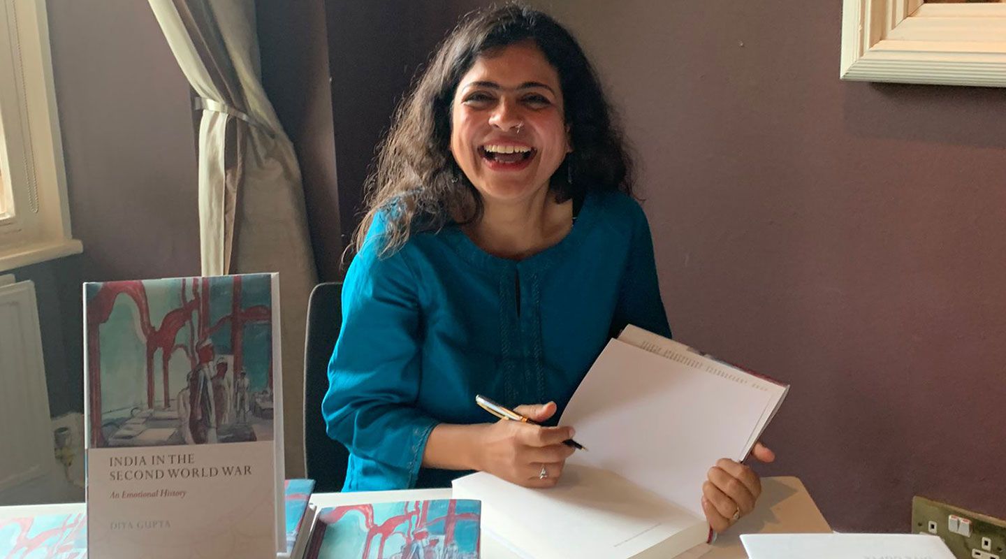 Dr Diya Gupta sits at a desk, wearing a bright turquoise shirt, and smiles as she signs a copy of her book.