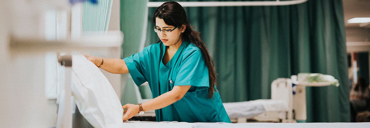 Young South Asian female nurse in a green uniform, making a bed in a hospital ward.