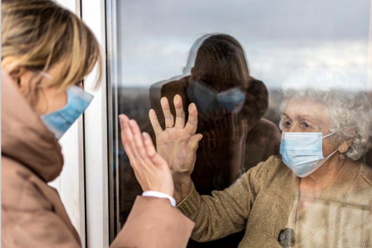 Elderly lady with a surgical mask on looking at a carer with a mask on standing outside her window.
