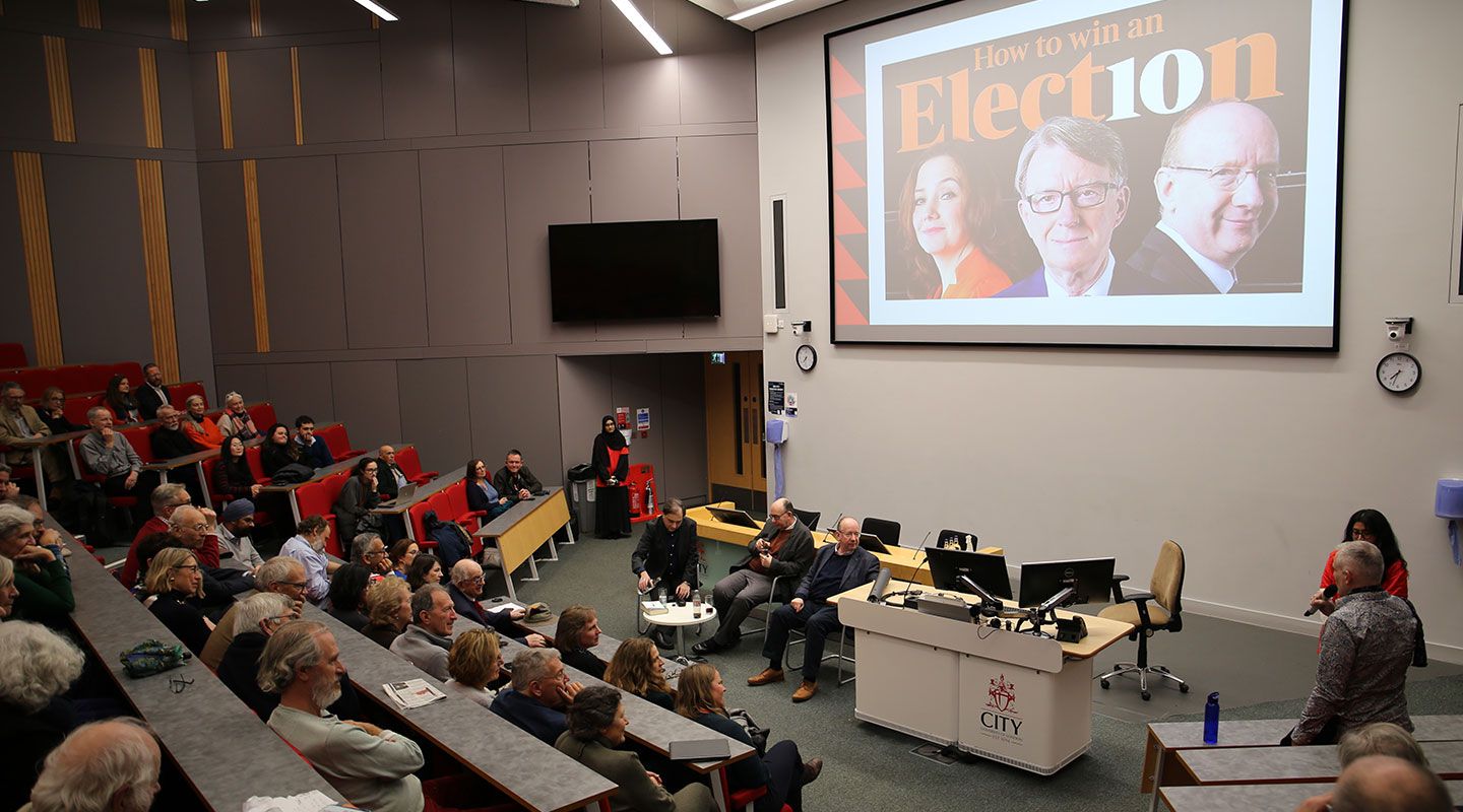 An audience member stands up and asks a question into a  microphone in a large amphitheatre full of people sitting on red seats. At the bottom of the image is Brett Spencer (Head of Centre for Podcasting Excellence), Prof Sir Anthony Finkelstein and Lord Daniel Finkelstein sitting on chairs and facing the crowd. Behind the speakers is a big screen with the thumbnail of Daniel's podcast 