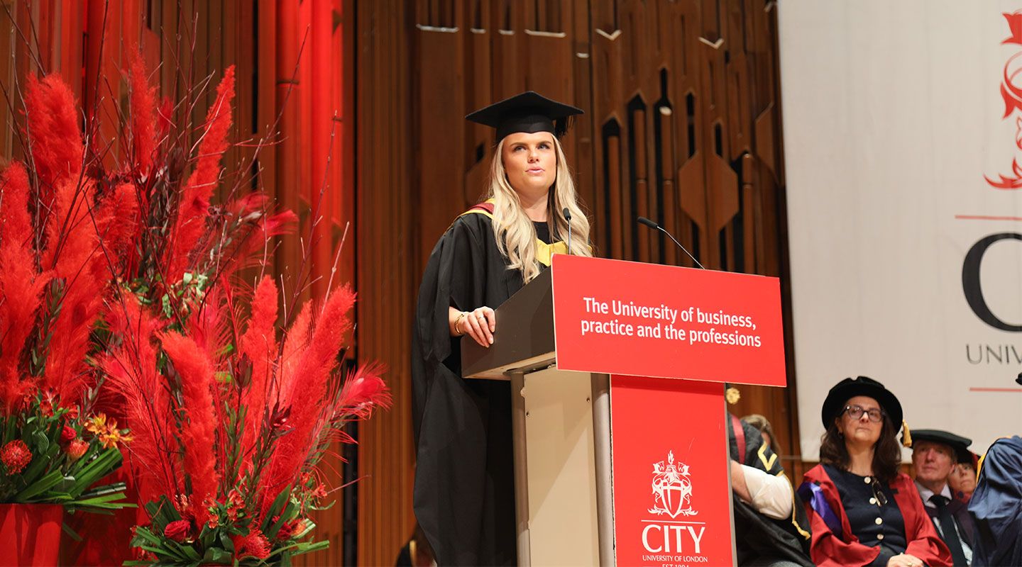 Student speaker Bibiane Beerens (MSc Corporate Finance) addresses the audience. She stands at the lectern at the centre of the image, which has City's slogan printed on it (The University of business, the professions, and practice). To her left is a large bouquet of red plants and flowers. To her right, academics sit in their formal robes on stage listening to her. She is in a hall in the Barbican Centre