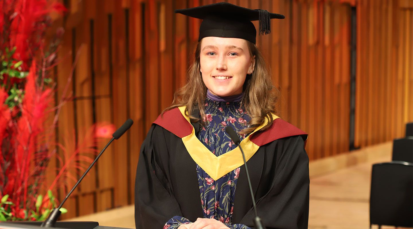 Picture of student speaker Gedvile Juodvalkyte standing in front of a lectern at the Barbican Centre. She wears a mortarboard and her graduation robe. To her left is a bouquet of  red plants. Behind her is the wooden structure of the Barbican building.