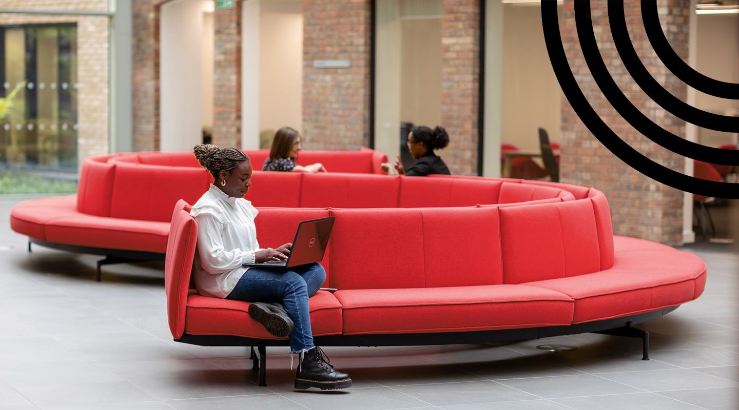 Young black woman using her laptop, sat on a bright red s-shaped sofa in a red-brick walled communal space with large windows.