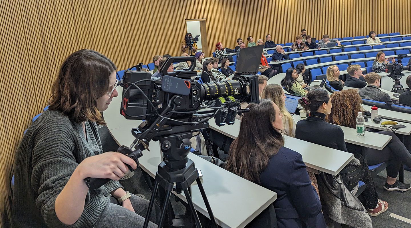 A student sits at a horseshoe lecture theatre desk holding a large filming camera. In front of her are rows of seats with other students