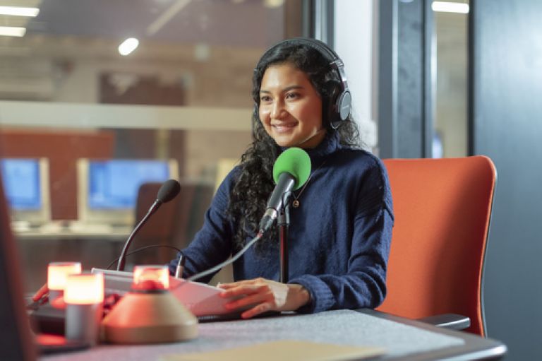 A young woman sits in front of a green microphone at an audio recording studio. A small red light is on, on the desk in front of her, signalling that she is currently recording. 