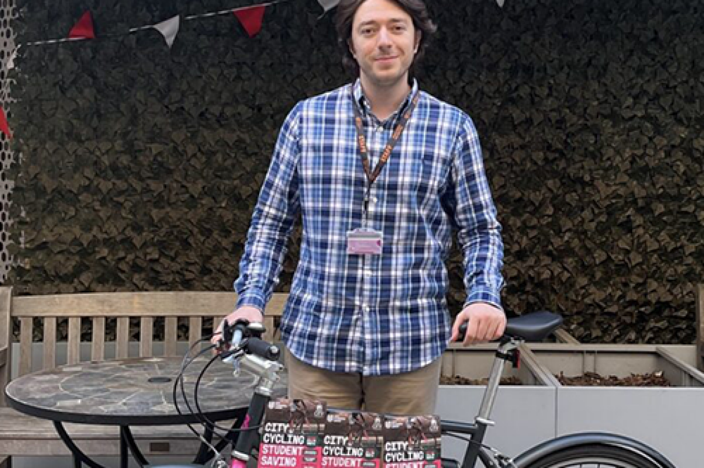 Yavuz Kafadar, Deputy President of City Student's Union, stands outside with a bike. He is wearing a blue shirt and beige trousers. There is colourful bunting in the background.