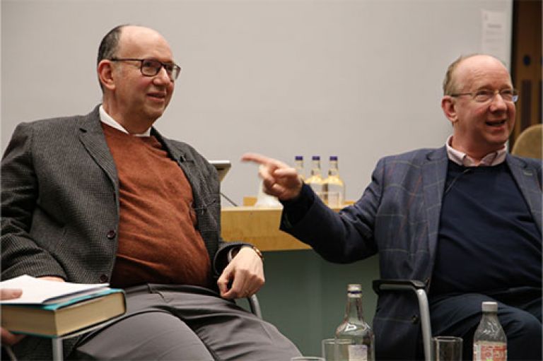 A picture of Prof Sir Anthony Finkelstein (left) and his brother Lord Daniel Finkelstein OBE (right). They are both sitting in chairs and smiling towards a crowd (not visible in the picture). Daniel is pointing at Anthony. On the lefthand-side, a book can be seen