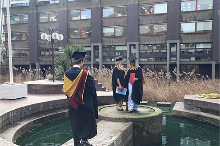 Image of two women wearing robes and mortarboards looking at each other and preparing to take a picture. They are standing ourtside at the Barbican Centre, surrounded by a semi-circle of water. In front of them, with his back to the camera, is a fellow graduate with his phone out, ready to take a picture of his friends. 