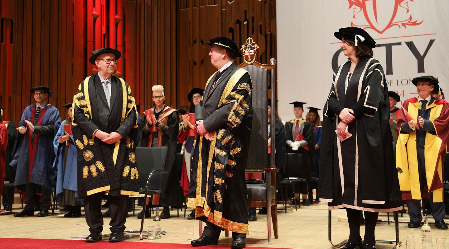 Picture of the Lord Mayor receiving his honorary doctorate and being made the Rector of City, University. He stands between Prof Sir Anthony Finkelstein and Dr Sionade Robinson. All three are wearing capes and hats, which are black with gold stitching. They stand on the stage at the Barbican in a hall. Behind them, academics sit and watch also in formal wear.