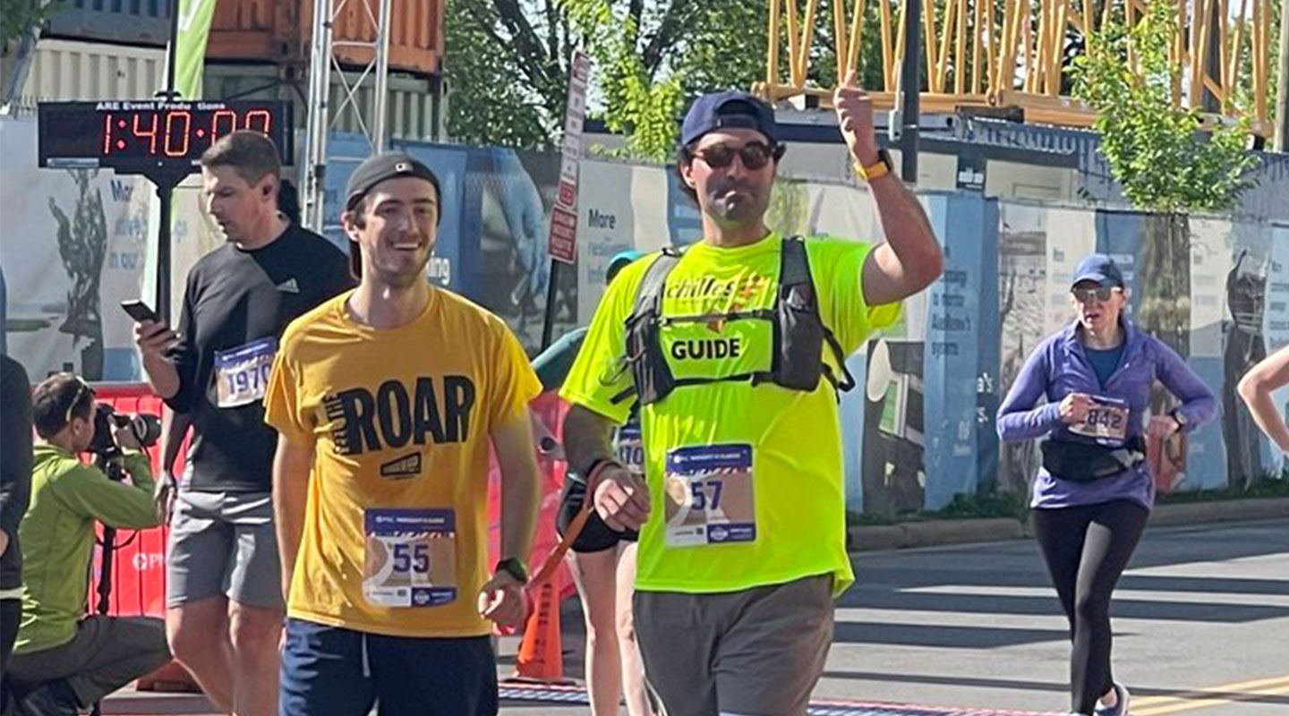 Tim Utzig begins a marathon alongside his sighted guide from Achilles International. He is smiling and wearing a yellow t-shirt which has his entrance number 55 stuck on it. To his right, his running guide gives a thumbs up and has the number 57 on his t-shirt. People run in the background.