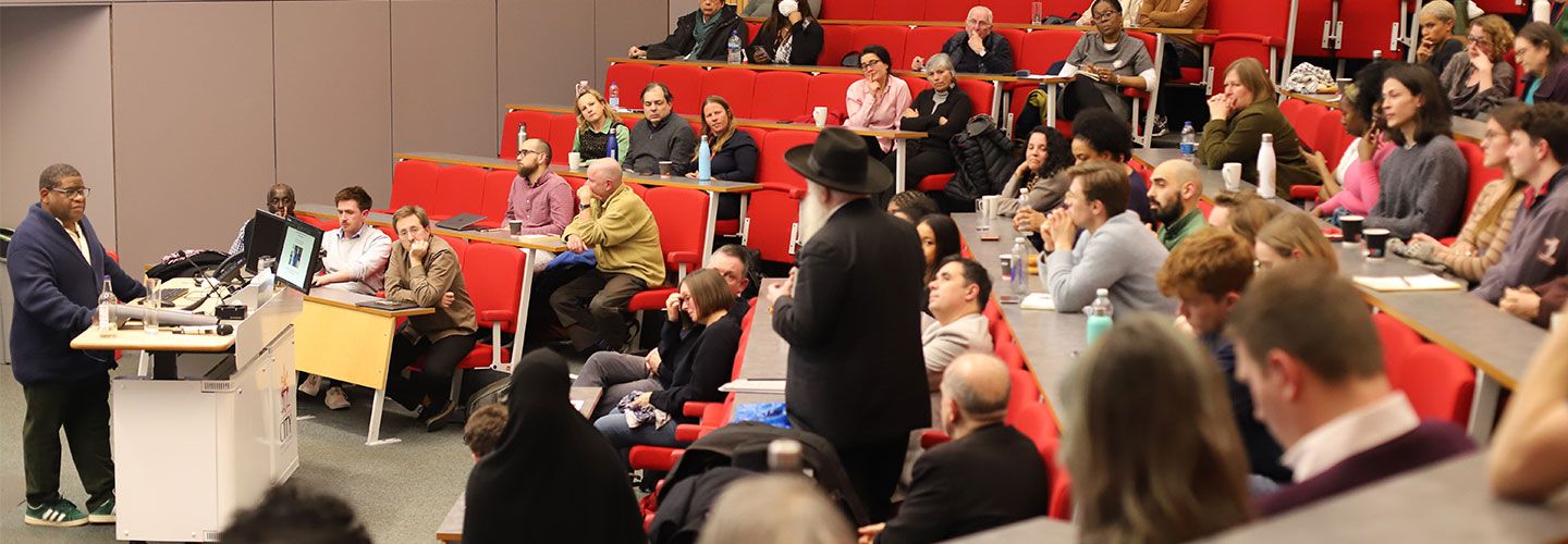 Prof Gary Younge stands in front of a desk. In front of him are rowing of desks, red chairs and stairs in an amphitheatre lecture hall. The seats are full of attendees. A man in the audience stands and asks a question