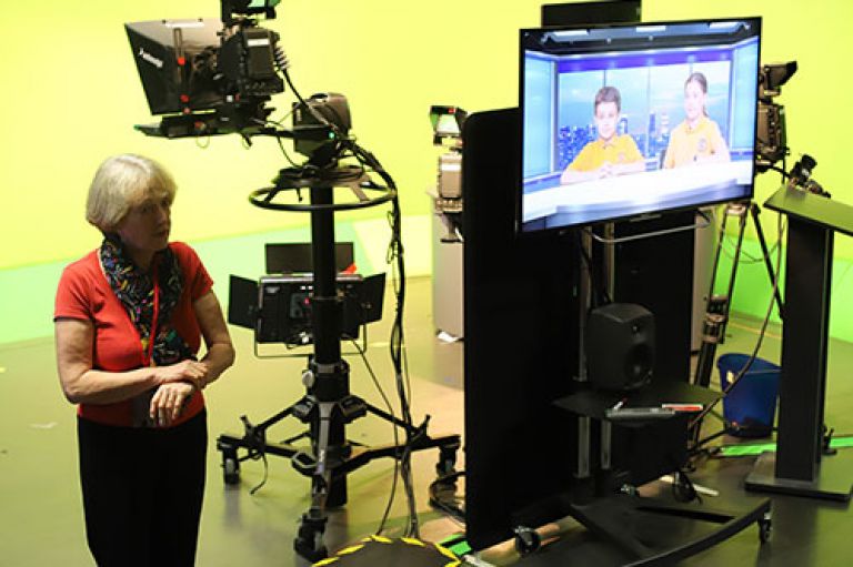 Prof Emerita Lis Howell stands in City's studio broadcast studio, which has a green screen, behind the camera. To her right is a screen which shows primary school two students sitting at a desk with a London skyline like they're on TV news. 