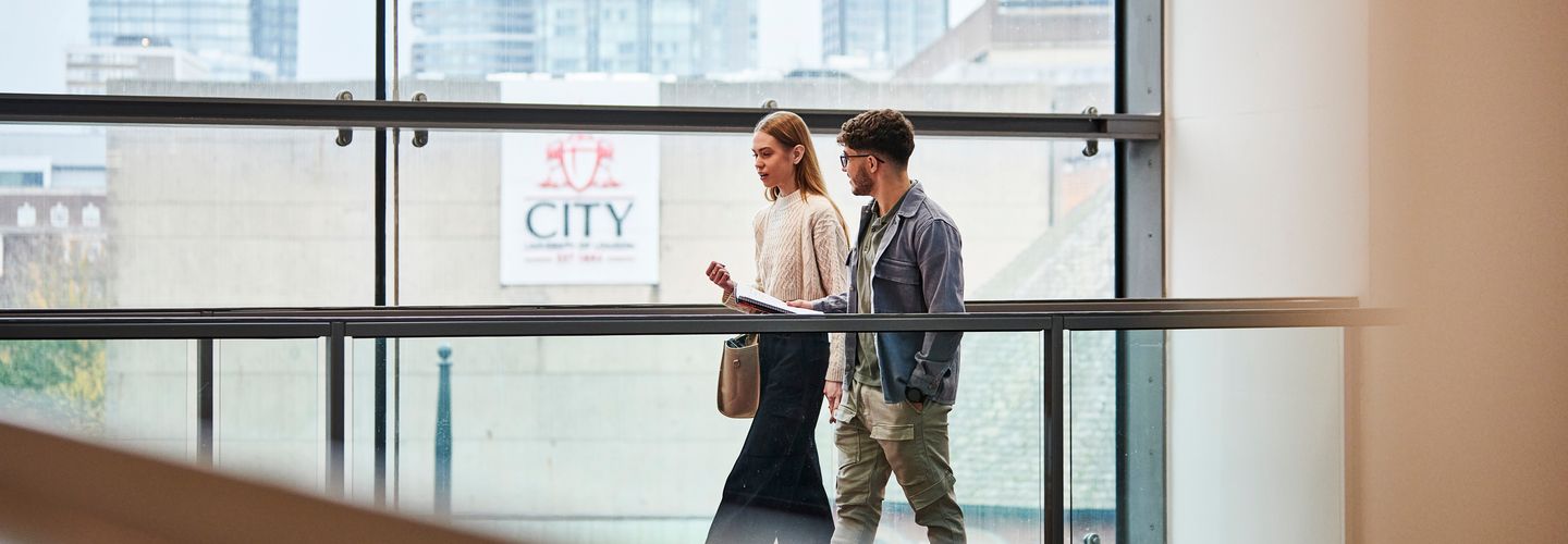 Two students, a man and a woman, walking along an upper walkway in Rhind Building, with a wide window view of the City behind.