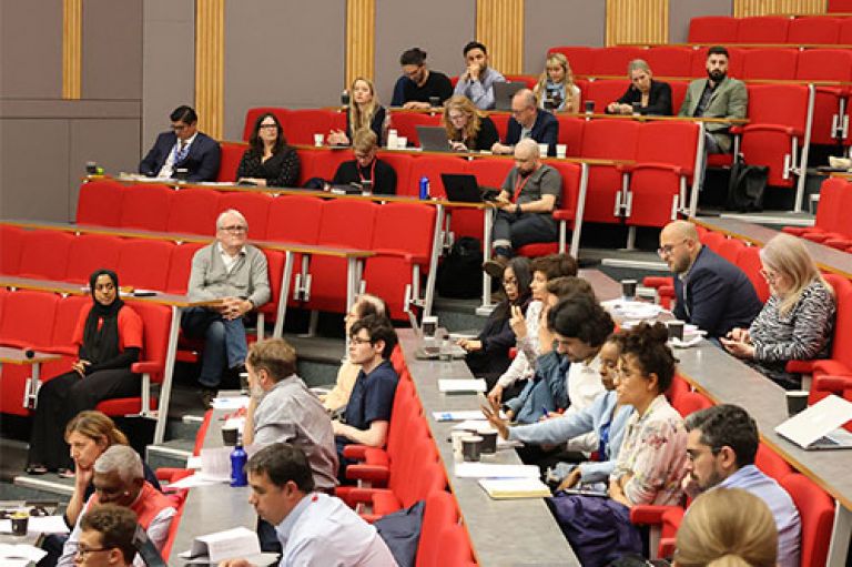 An image of rows of attendees sitting on red chairs with desks in front of them, some with notebooks, laptops and phones, in an amphitheatre-style room.