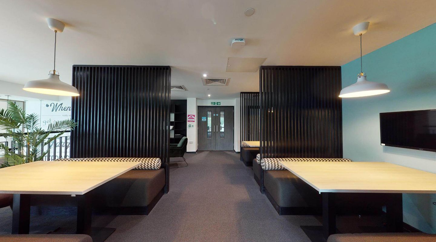 Modern communal space with large tables and cushioned bench seating with black slatted dividers, and screens on the wall
