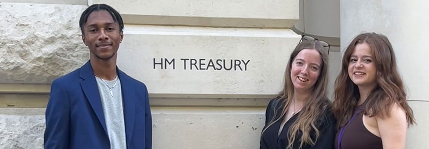 Student Miles Johnson stands on one side of the words 'HM Treasury' printed against a building, and to the other side stand two fellow students.
