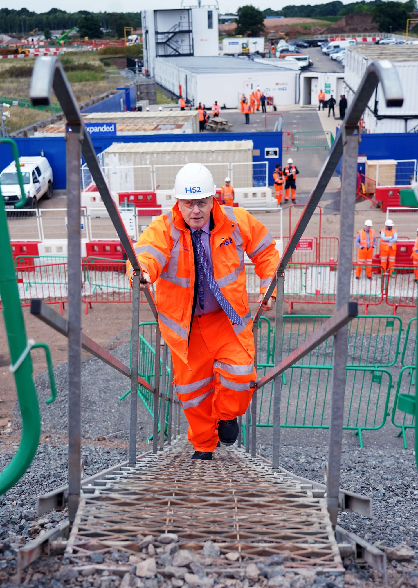 An image of former Prime Minister Boris Johnson in bright orange construction clothing climbing up a metal stairway. Behind him is a construction site.