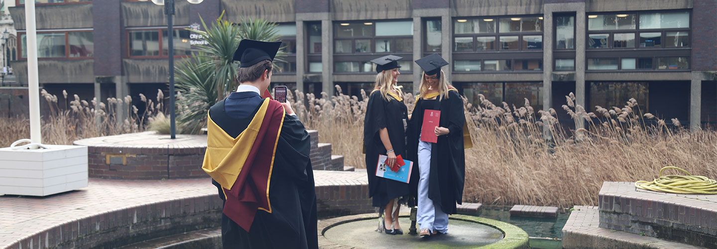 Image of two women wearing robes and mortarboards looking at each other and preparing to take a picture. They are standing ourtside at the Barbican Centre, surrounded by a semi-circle of water. In front of them, with his back to the camera, is a fellow graduate with his phone out, ready to take a picture of his friends.