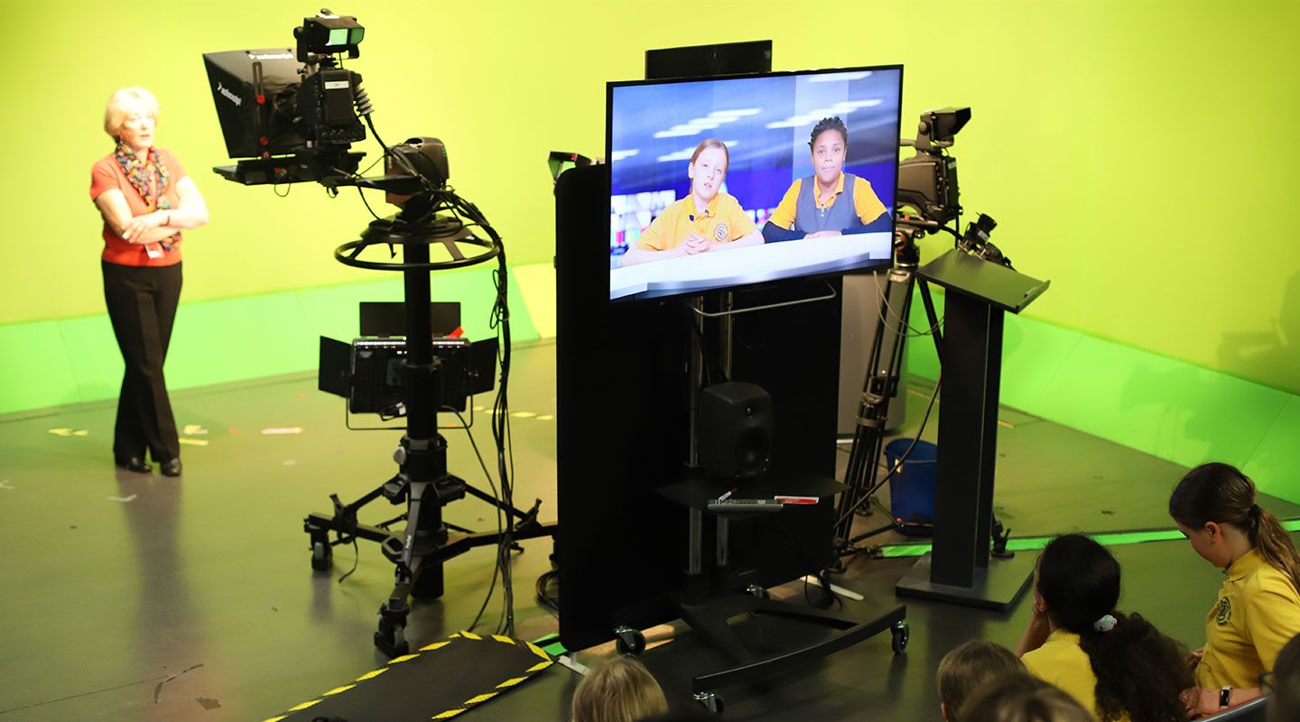 Prof Emerita Lis Howell stands in City's studio broadcast studio, which has a green screen, behind the camera. To her right is a screen which shows primary school two students sitting at a desk with a London skyline like they're on TV news.