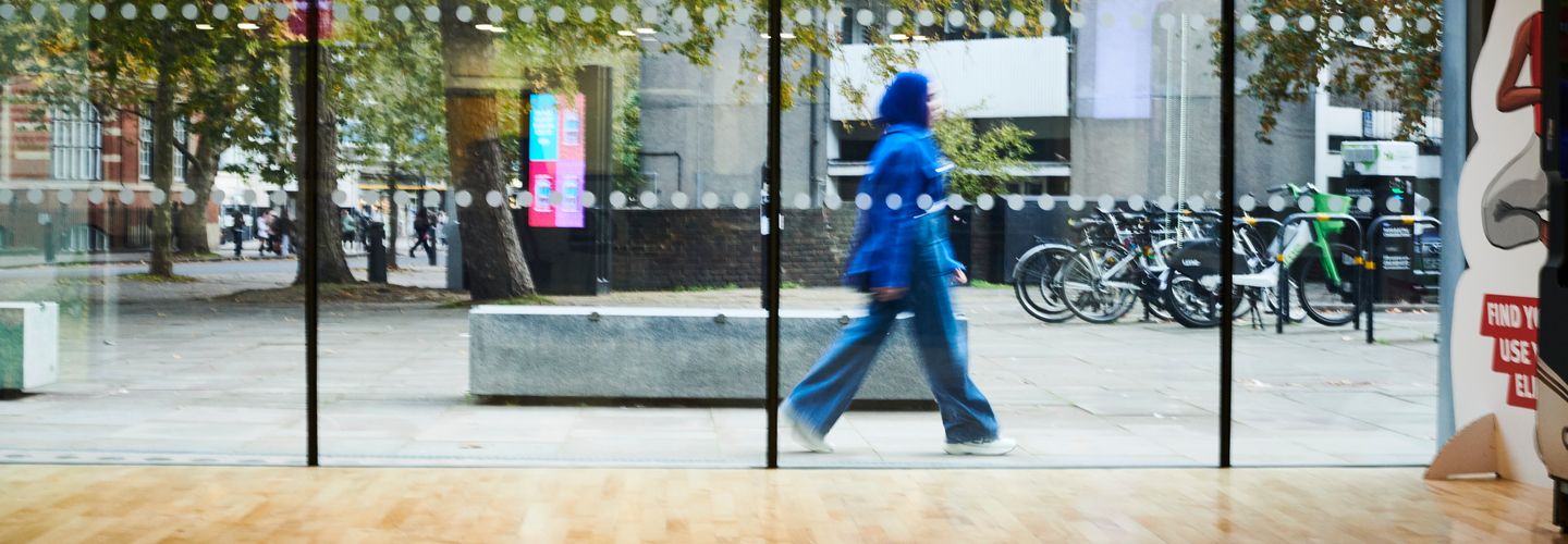 A young woman in a bright blue coat and headscarf walks past the entrance of Rhind Building, photo taken from inside looking out.