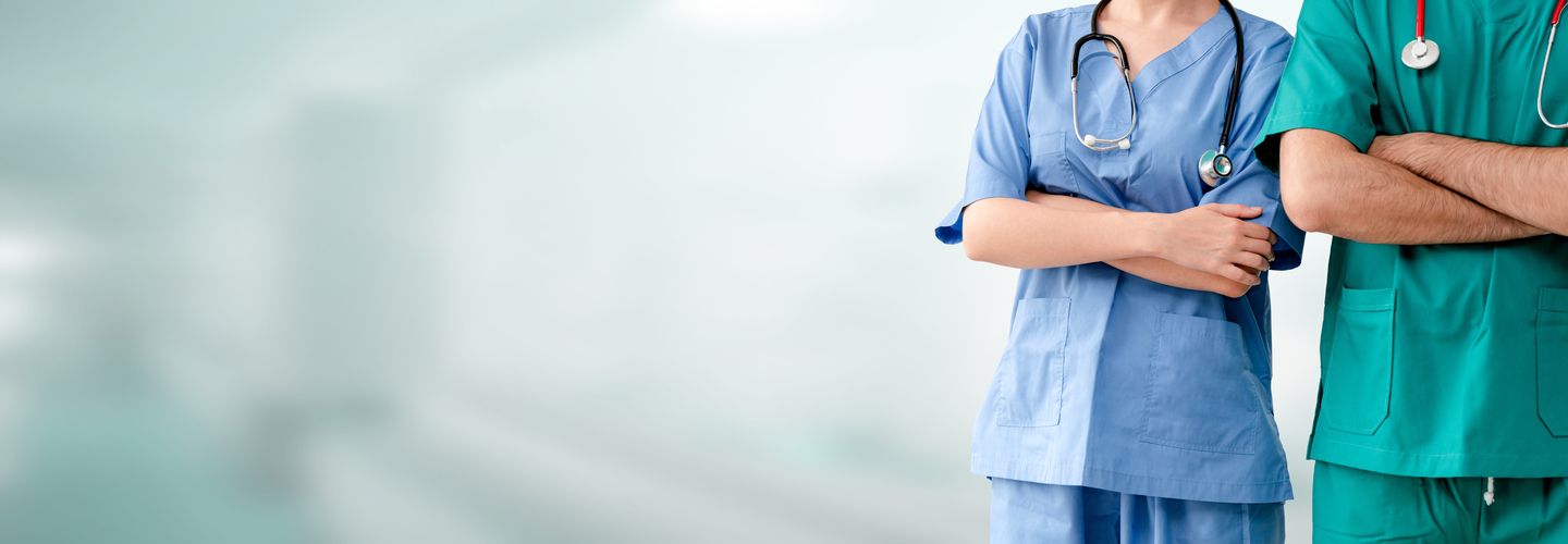 Torso view of male and female doctors in scrubs, standing side by side with arms crossed and stethoscopes over their shoulders..
