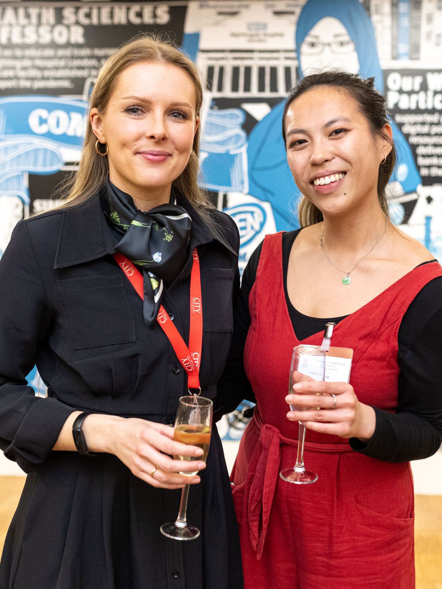 Picture Xenia Kotova (left) and Meghan Yang (right) at the City Professional Mentoring Scheme awards. They are holding a glass of fizz and are standing in front of a backdrop of a wall covered in cartoon drawings.