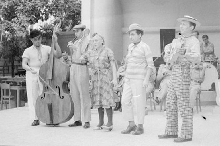 A black and white picture of five people dressed up smartly on a stafe, with musicians sitting on seat behind them. the firts person is a man wearing a hat and playing a cello. The second person is a man wearing a flatcap playing a flute. The third is a woman in a dress holding a percussive instryment. the fourth person is wearing a striped top and hat. the final person is playing a guitar.