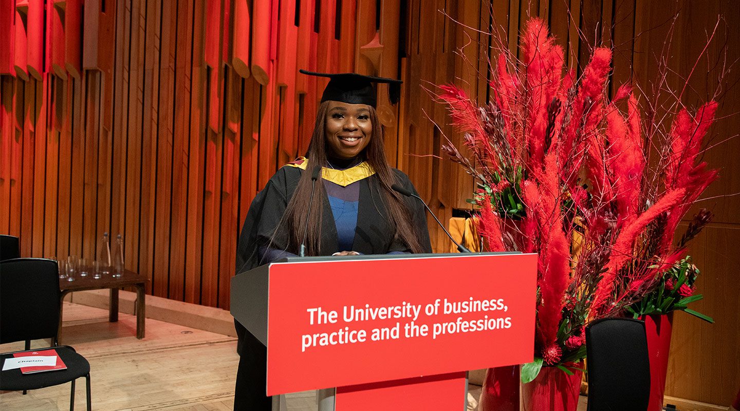 Portrait of graduate speaker Inemesit standing in front of the lectern which has the words of City's slogan printed across it (The University of business, practice and the professions). Behind her, to her right, is a large bouquet of red flowers and plants. She is standing in a lecture hall in the Barbican and smiling to the camera, wearing her gown and mortarboard.